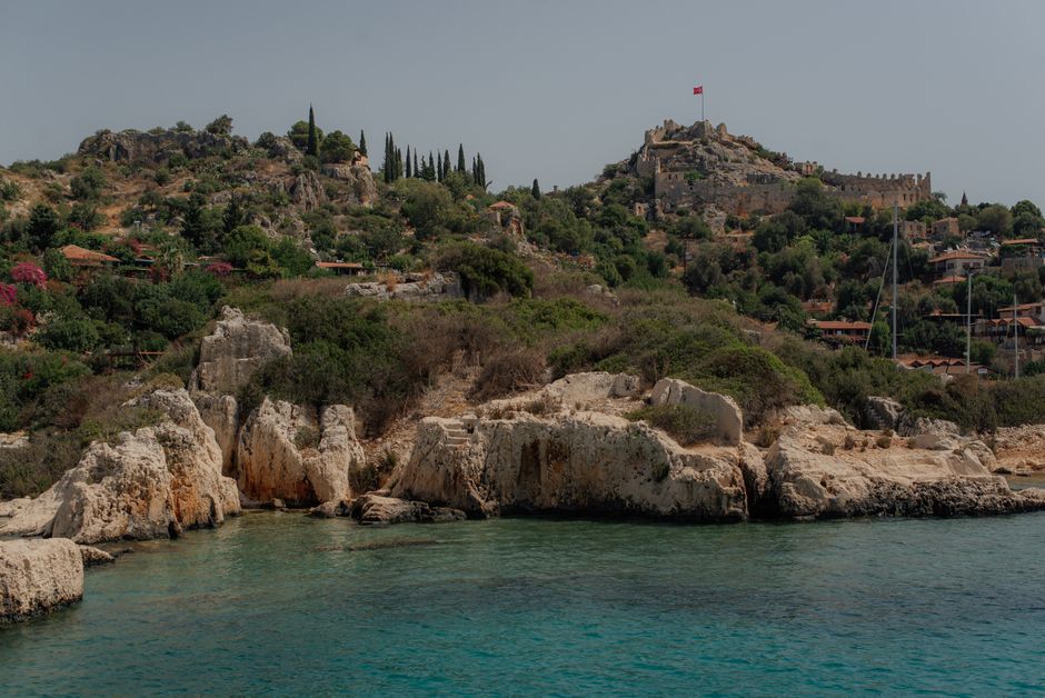 A picturesque view of the Mediterranean coastline featuring rocky shores and historic hillside ruins under a clear sky.